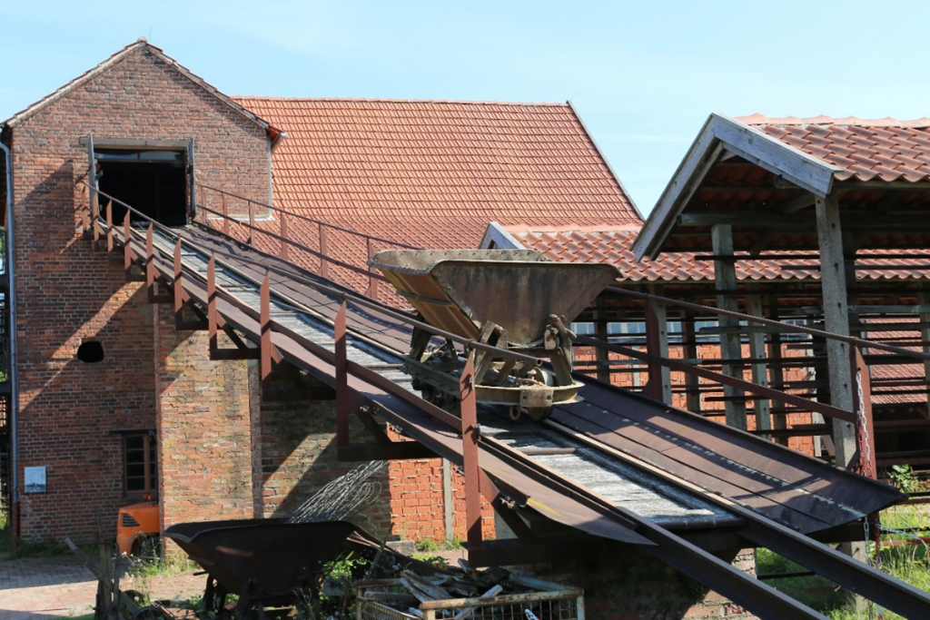 Eine schräg nach oben verlaufender, schienengebundener Schrägaufzug mit einer Kipplore darauf. Der Schrägaufzug führt in das Obergeschoss eines zweistöckigen, aus roten Ziegeln gemauerten Gebäudes.