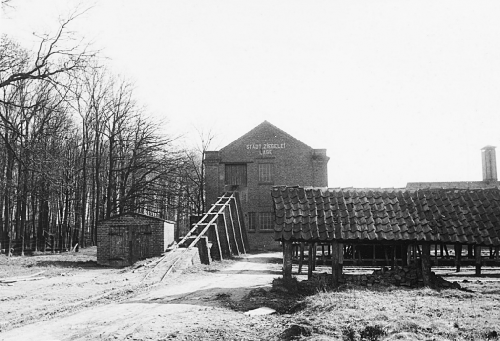 Im Vordergrund ein langgestrecktes, offenes Holzgebäude mit einem Ziegeldach, das auf Holzpfosten ruht. Dahinter ein schienengeführter Schrägaufzug für Kipploren, der in das Obergeschoss eines zweigeschossigen Backsteingebäudes führt. Auf der Fassade des Gebäudes steht in großen Buchstaben 'STÄDT. ZIEGELEI LAGE'. Links neben dem Schrägaufzug befindet sich ein kleineres, ebenfalls aus Backstein gebautes Gebäude, das als Lokschuppen dient.